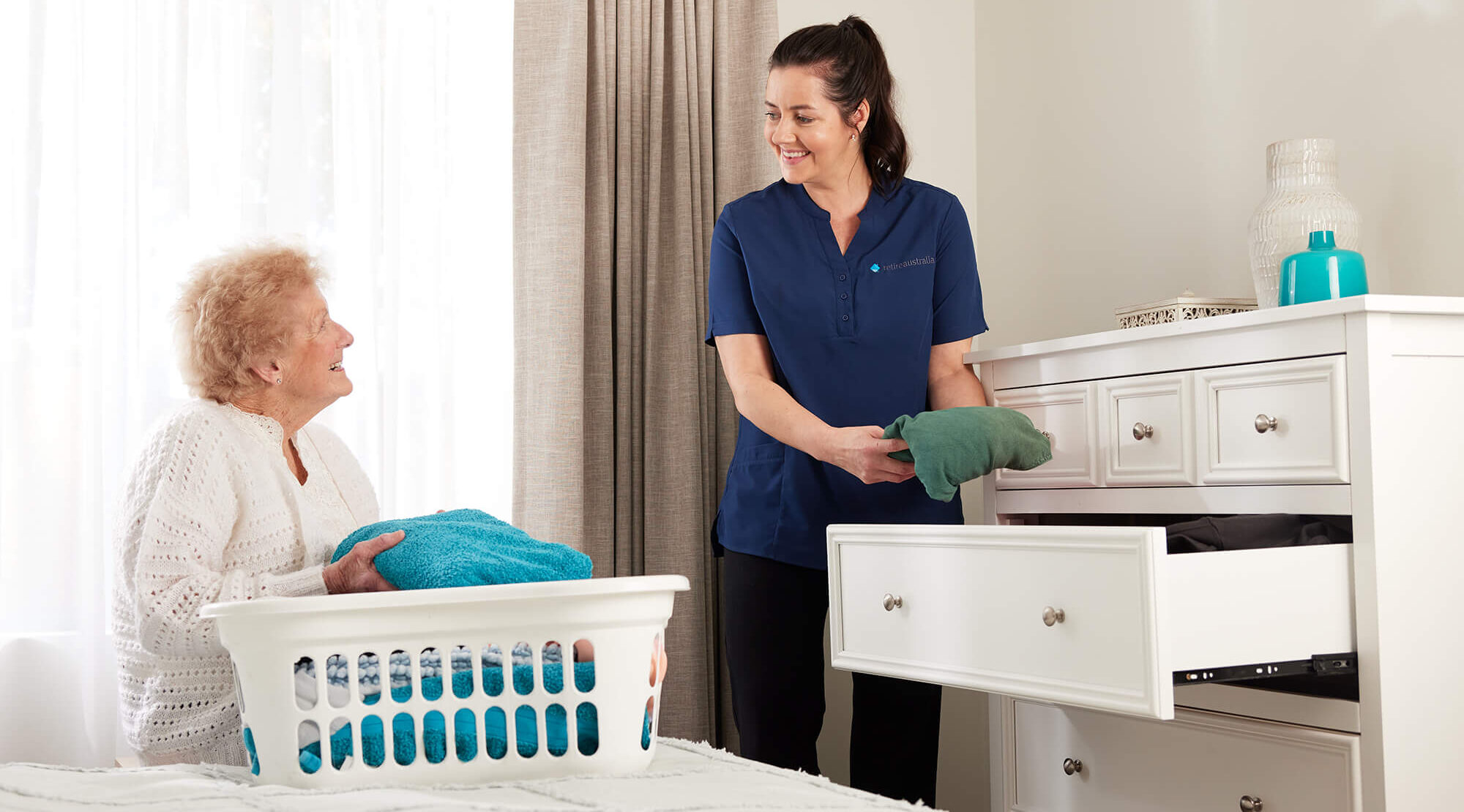 Resident sitting on bed as RetireAustralia employee assists with putting away clothes in the dresser