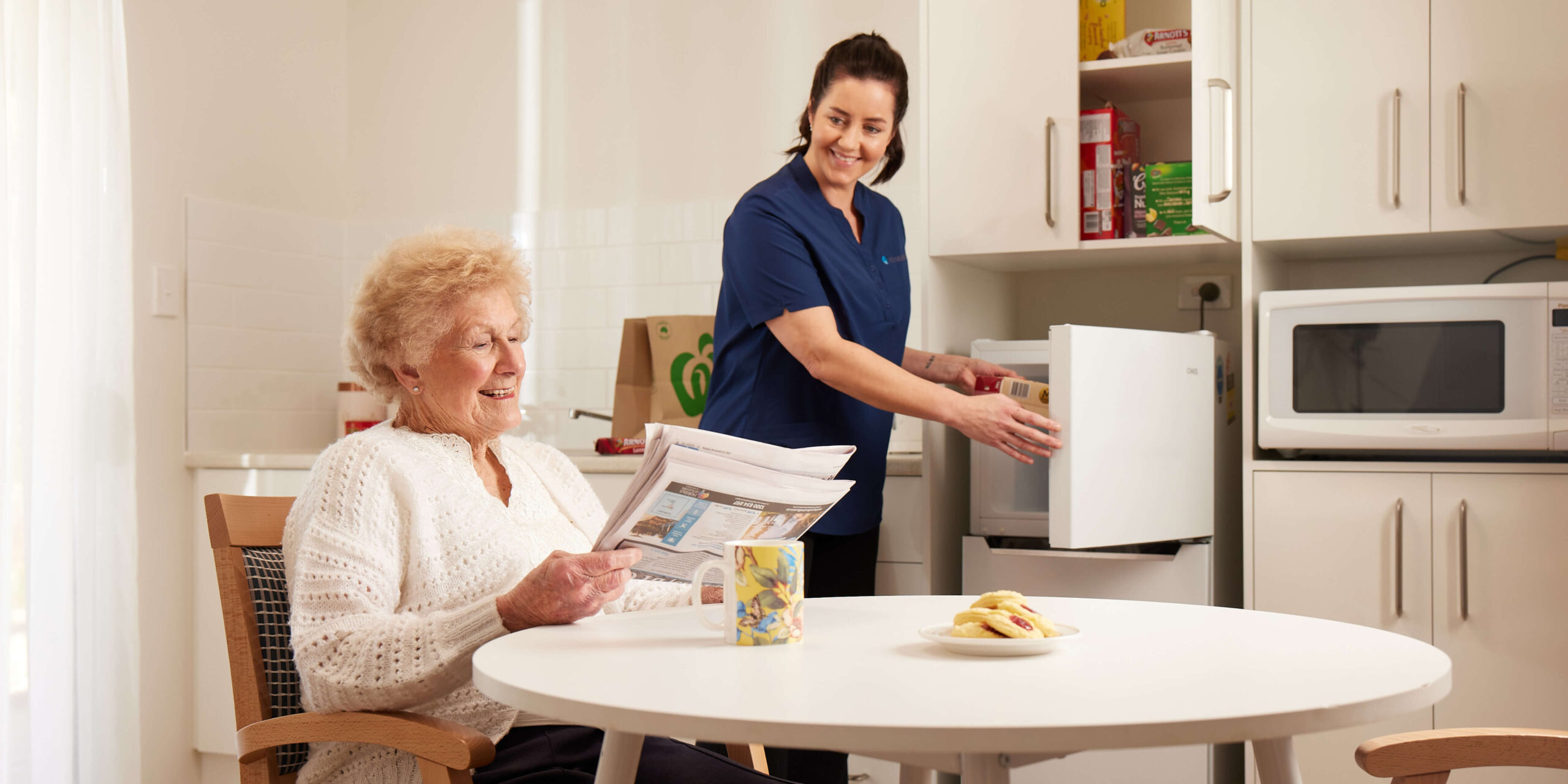 Resident reading her morning paper whilst RetireAustralia employee assists her by putting away her food.