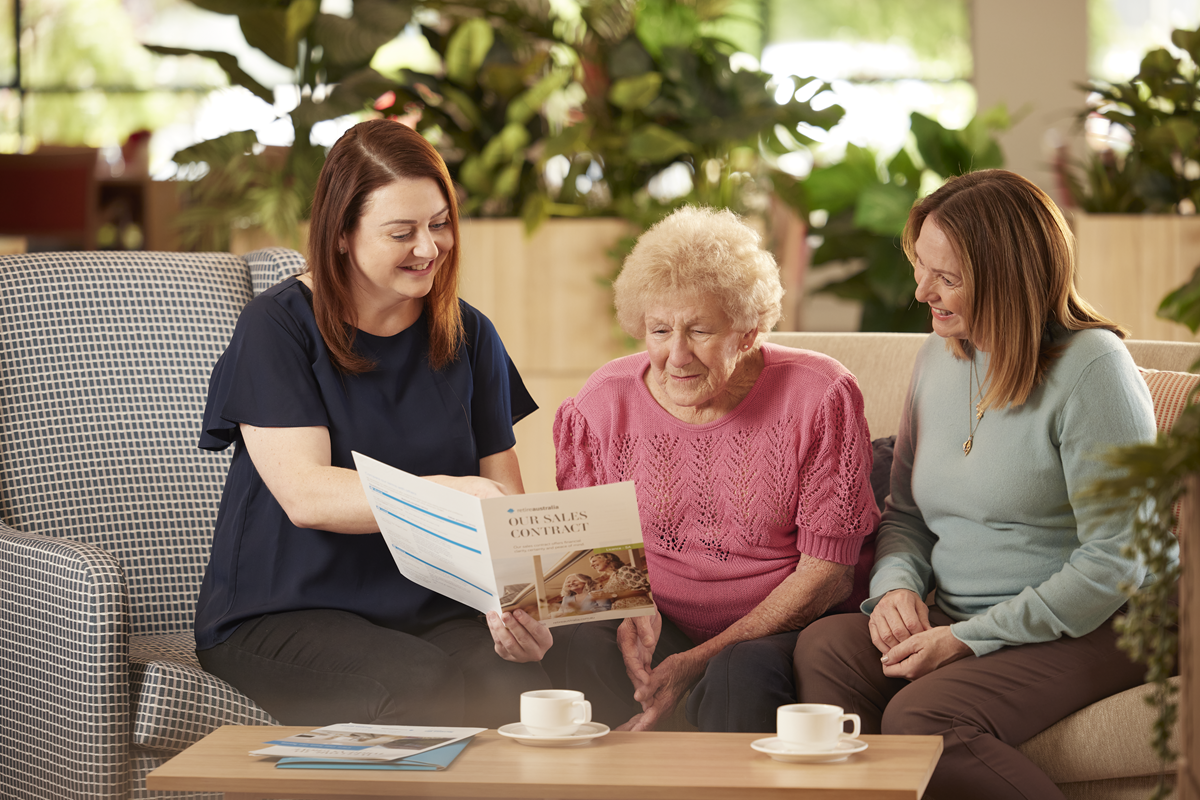 Sales consultant in a dark blue top shows an older lady and her younger daughter a sales contract brouchure at a retirement village sales appointment