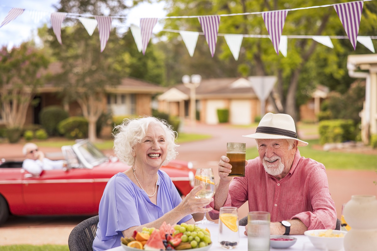 An elderly couple sitting outdoors at a table, smiling and raising their drinks in a toast.