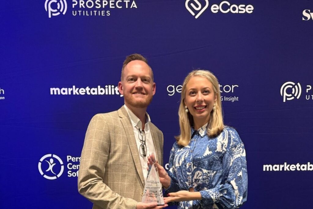 Man and woman standing in front of a blue media wall holding an award and smiling. The background displays logos including "Prospecta Utilities", "eCase", and "marketability"